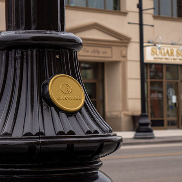 A KSB16 Florentine Sr. decorative split base accompanied by a custom 'Oakville' emblem on a fluted pole. This project is on one of the busy streets of Oakville, Ontario.